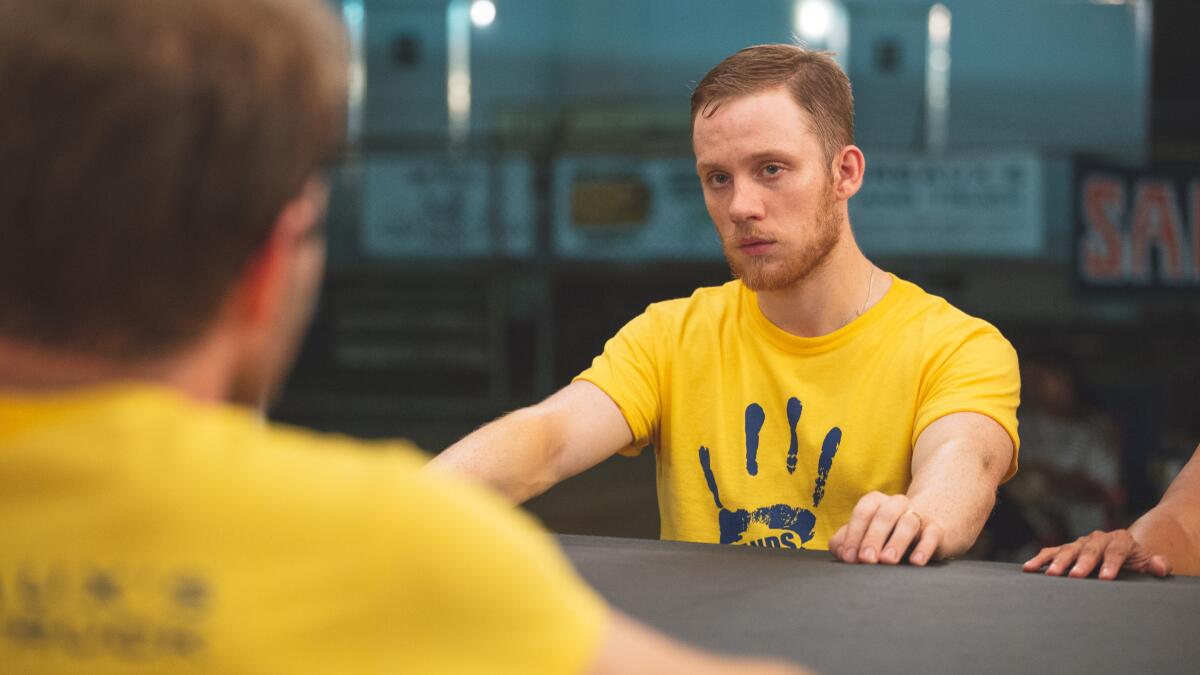 A bearded man in a yellow T-shirt sits with his hands resting on a table, looking hostlie.