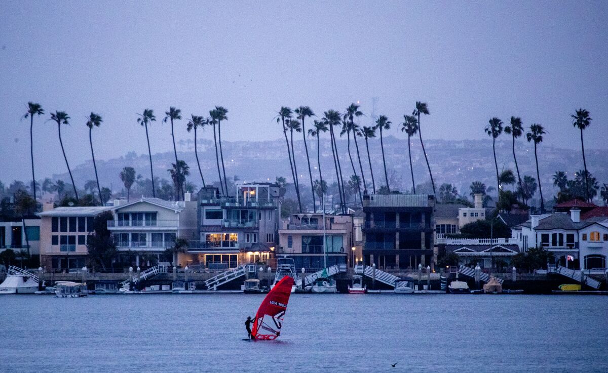 A windsurfer sails on the ocean in front of buildings and palm trees at dusk.