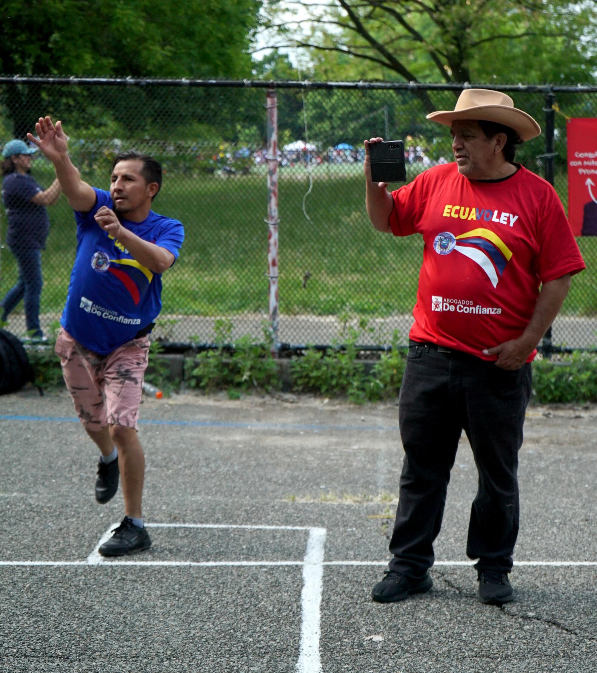 A man wearing blue plays ecuavoley while another wearing red and a cowboy hat films him.