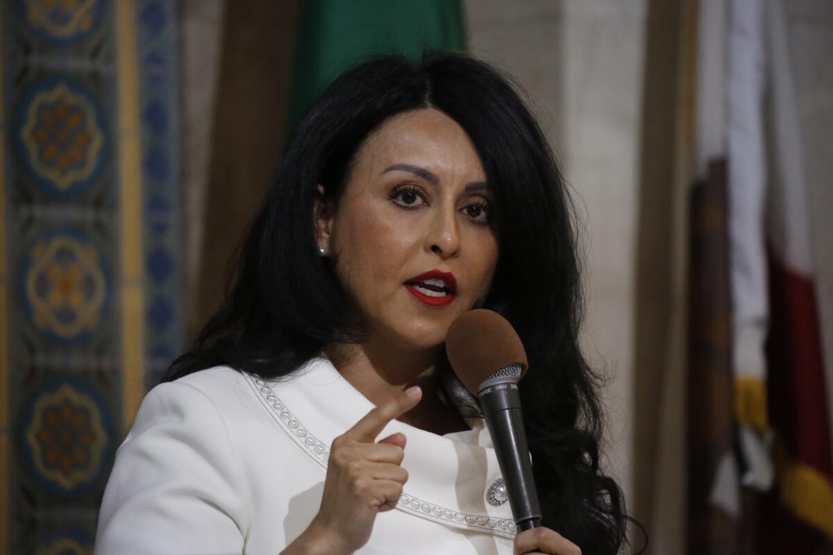 A woman with long dark hair and wearing a white suit talks into a microphone. 