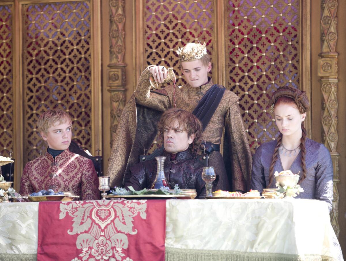 A man wearing a crown pours drink on the head of a man sitting at a table between a young girl and boy.