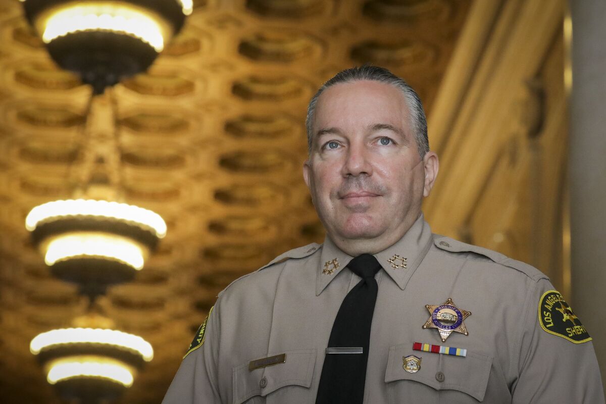 A man in a khaki-colored uniform with black tie and a sheriff's badge against a backdrop of lights