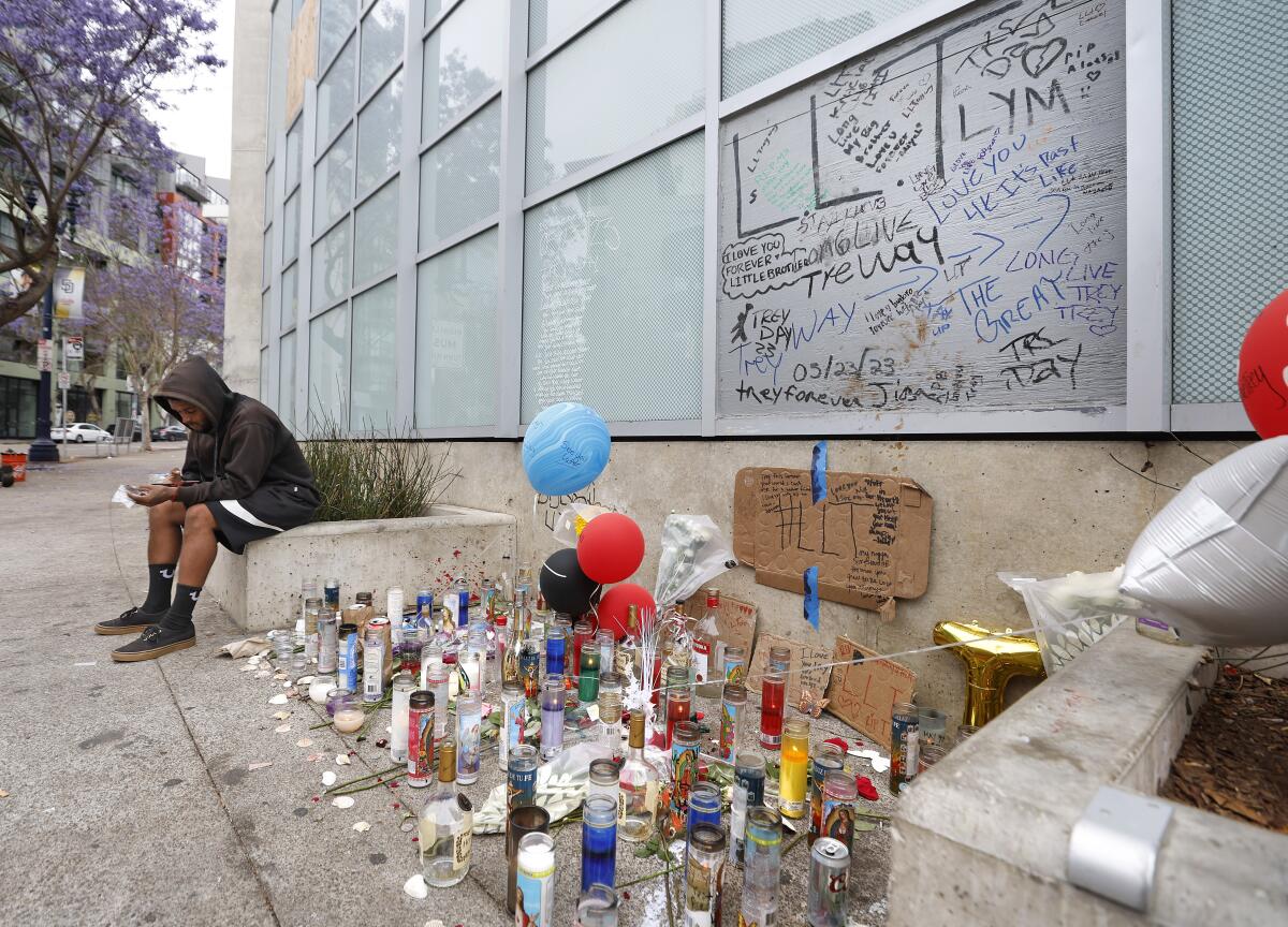 A person in a hoodie sits near a memorial of candles, balloons and notes outside a building.