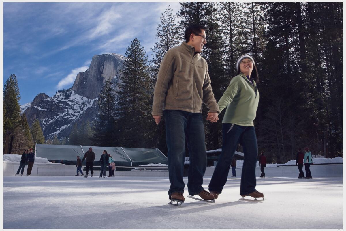 A couple holds hands ice skating in front of trees and mountains.