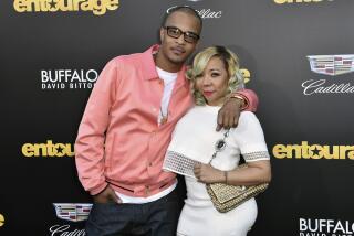 T.I. in a pink satin jacket posing with his right hand in his pants pocket. His left arm is around Tiny in a white dress