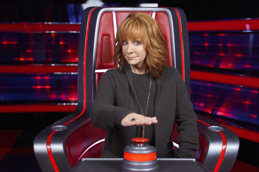 Reba McEntire wearing a dark outfit sitting on a red chair with her right hand hovering over a red button 