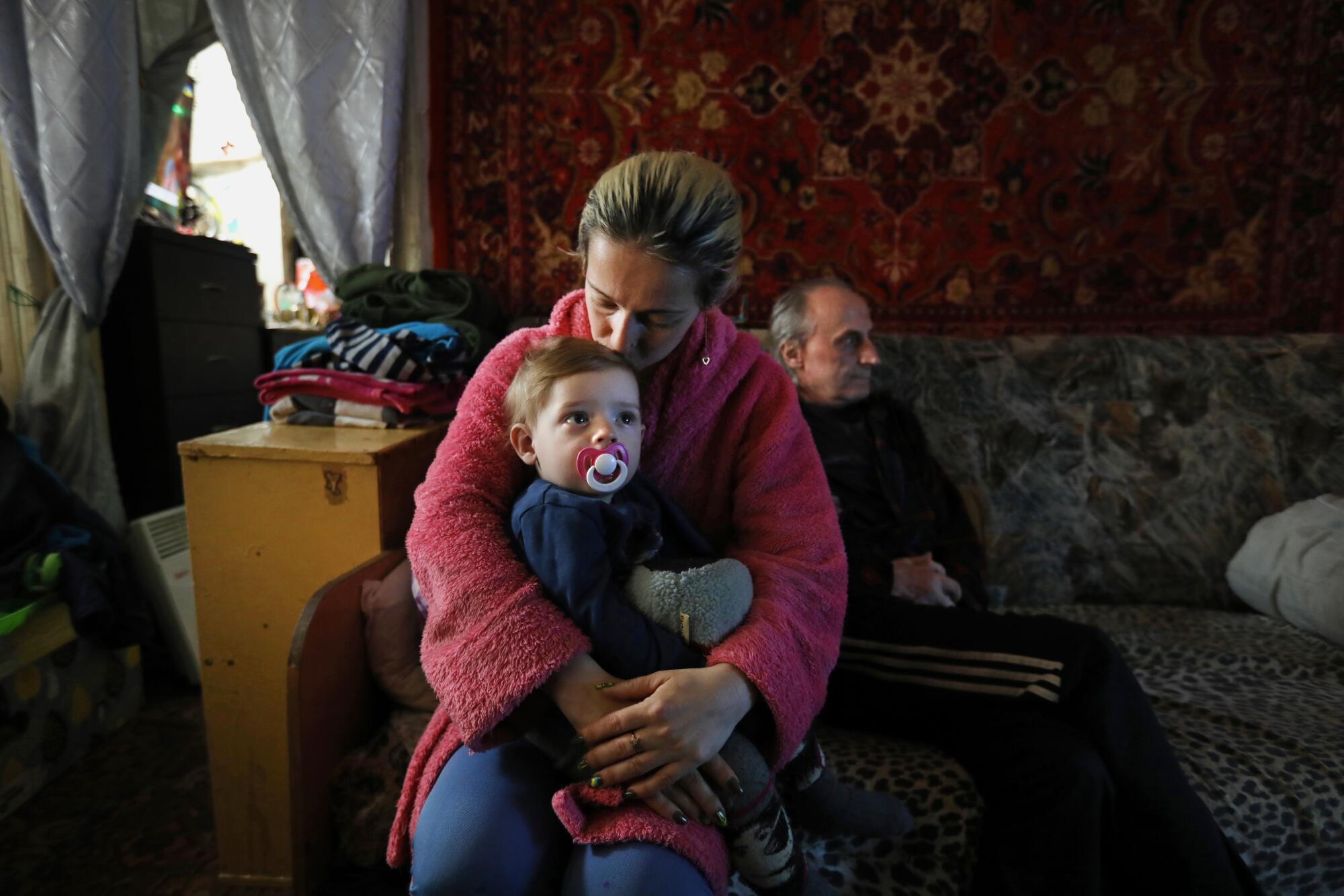 A woman kisses her baby while a man sits behind them in a small apartment.