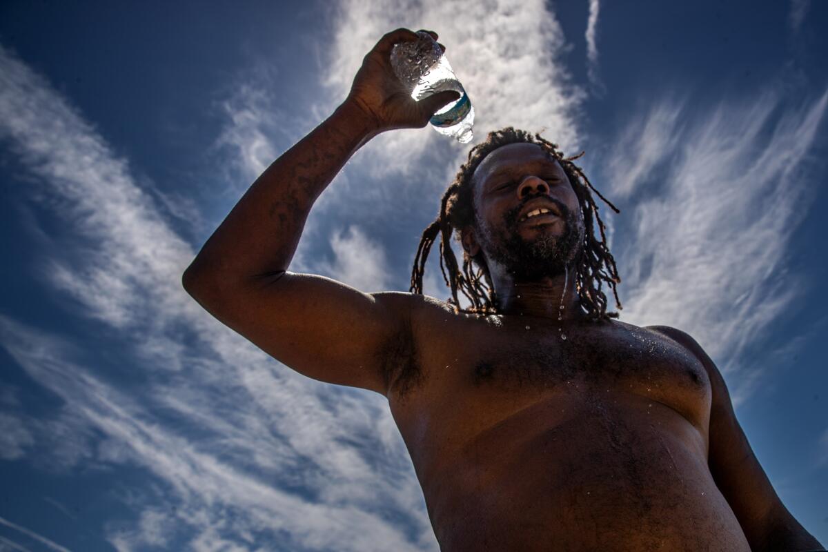 A shirtless man stands outdoors and pours water on his head in an attempt to cool off.