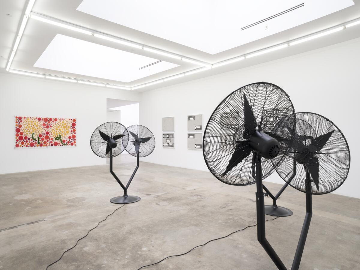 An installation view of an art exhibit in a gallery shows large fans on the floor and a painting made of colorful dots