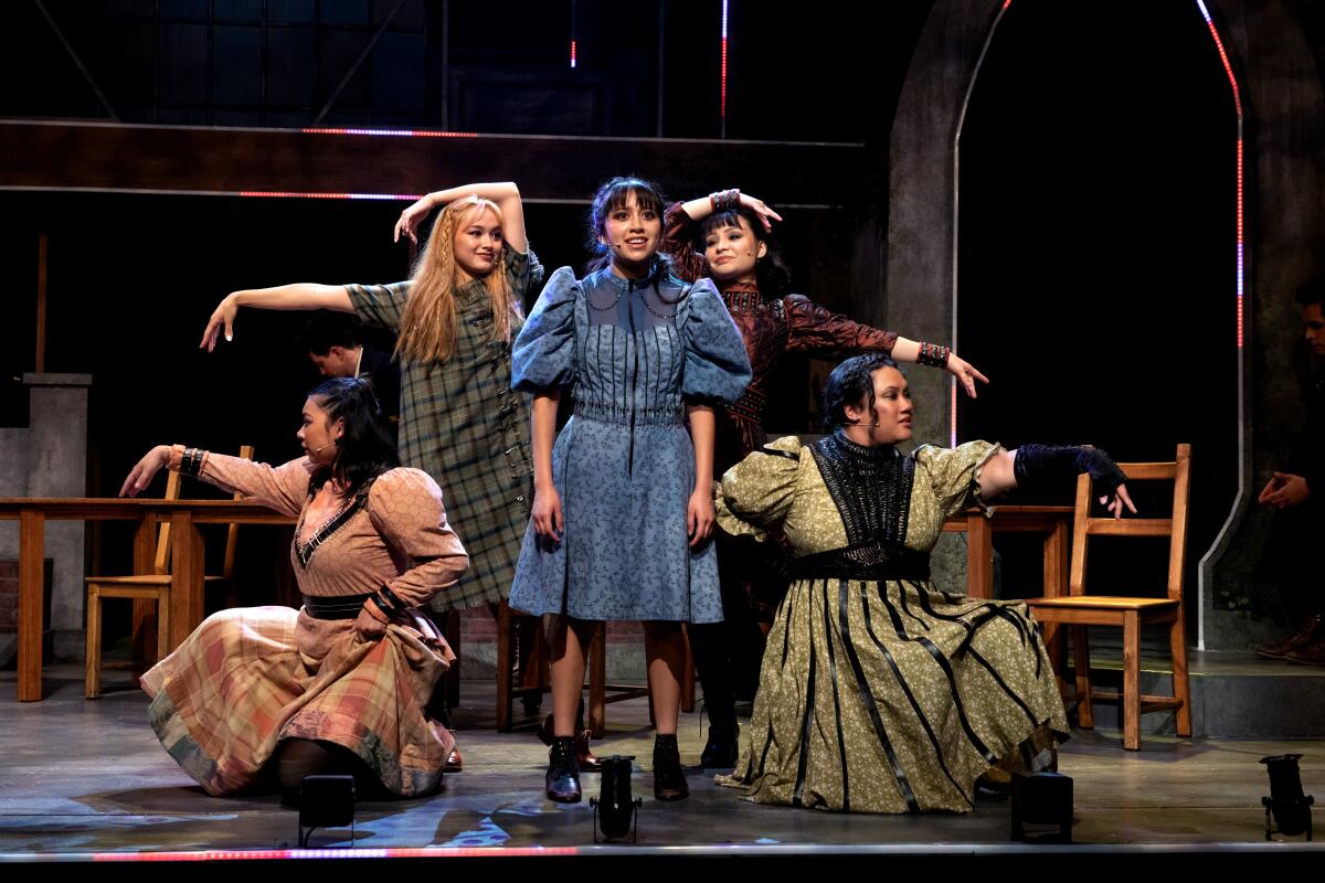 Four young women stand around a fifth, posing in 19th century costumes onstage