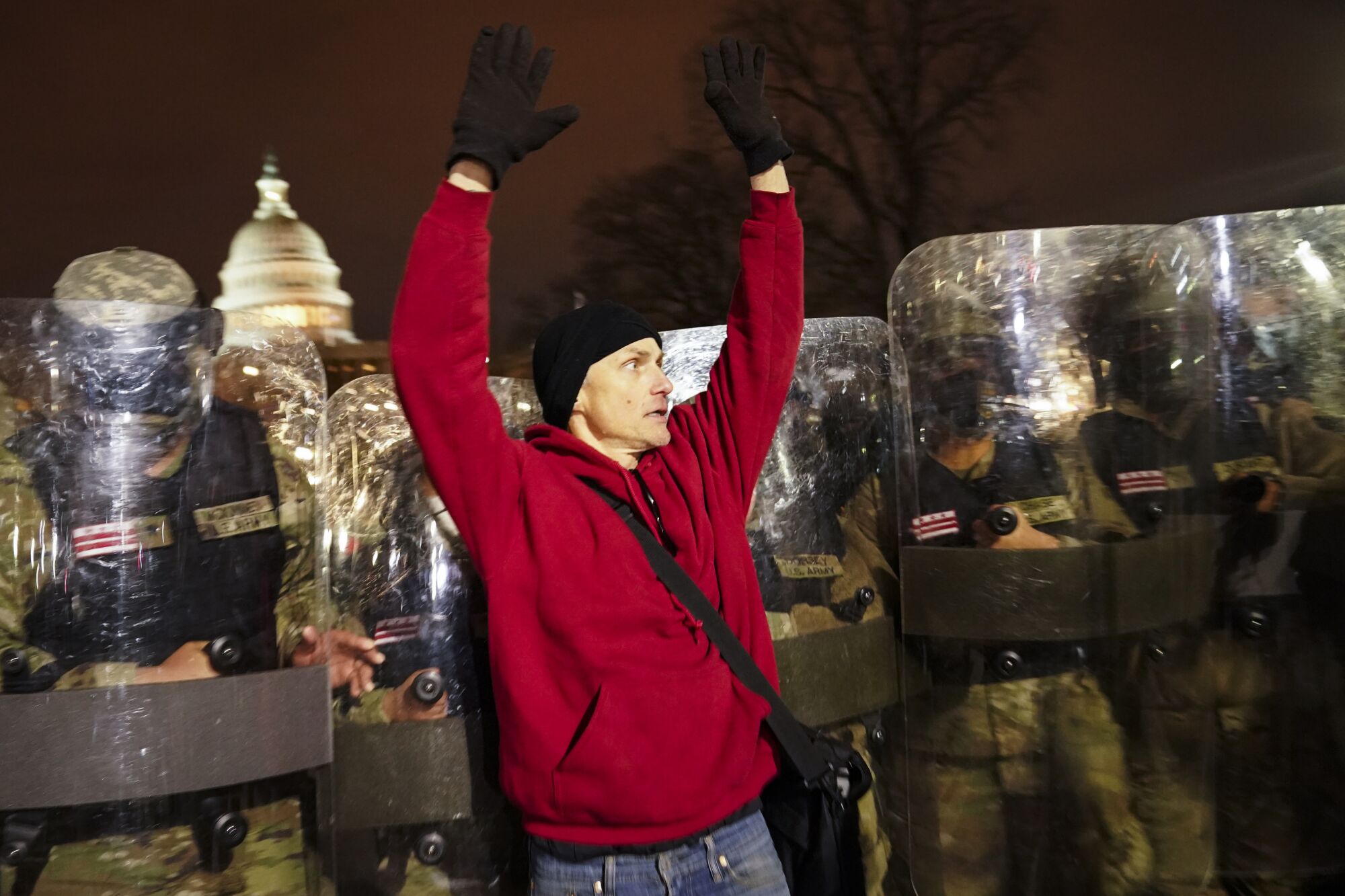 A man in a red sweatshirt holds up both arms as he's pushed along by a line of National Guard members in riot gear.