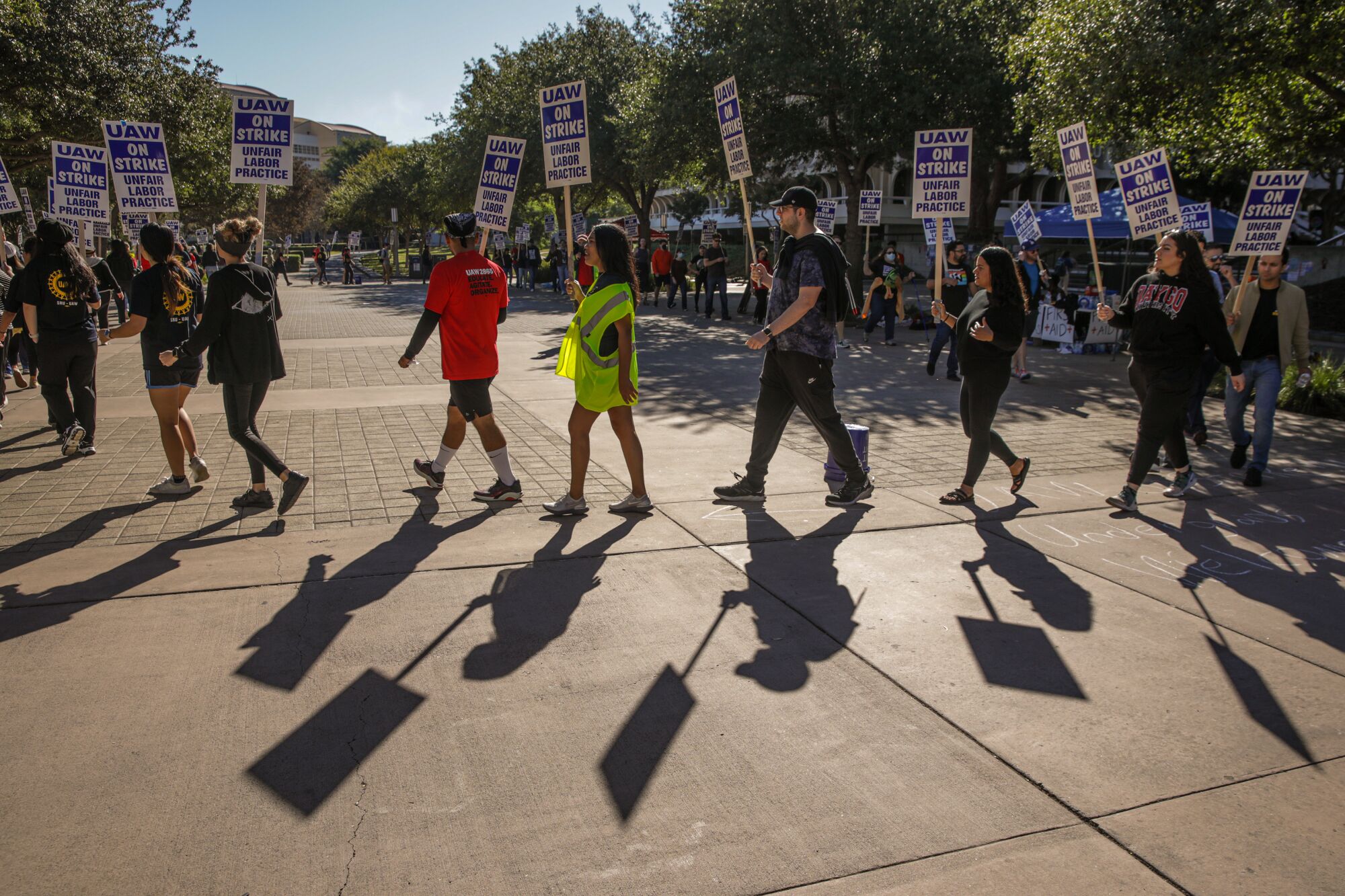 A group of demonstrators hold signs and walk in a large circle while they picket at University of California Irvine.