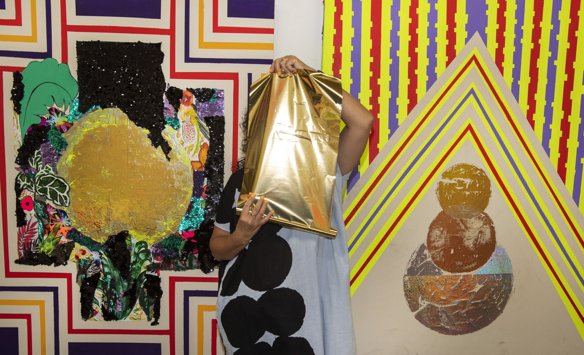 Carolyn Castaño, in a gray and black dress, holds up a gold sheet in front of her face as she stands before paintings