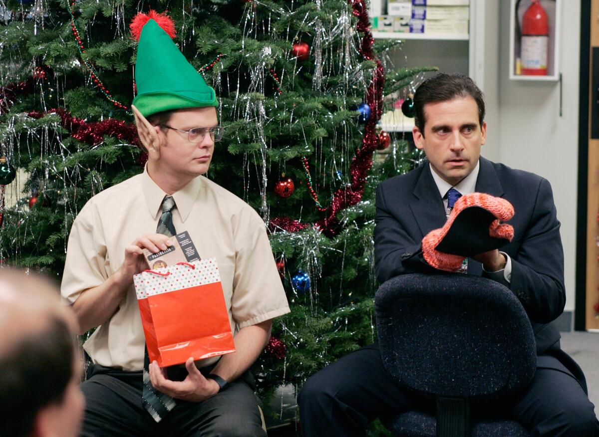 A man wearing an elf hat and another man sit in front of a Christmas tree in a holiday episode of "The Office"