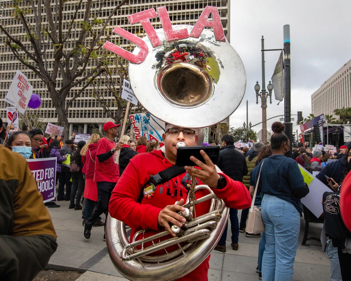 A young man in glasses and a red sweatshirt plays the tuba as a crowd gathers behind him.