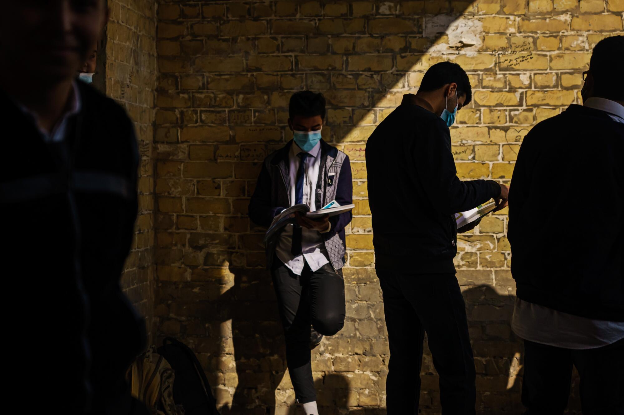 Young masked men in school uniform hold books as they stand near a brick wall 