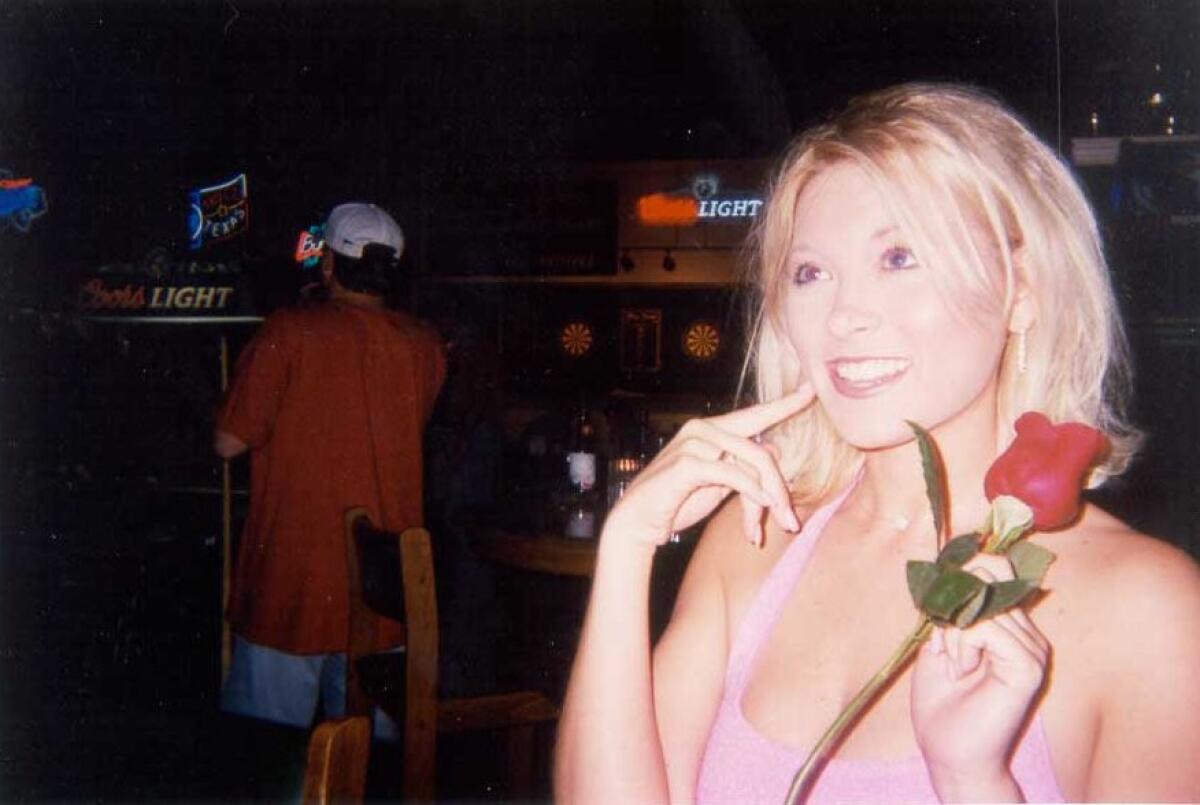Amy Carlson in a pink tank top holds a rose in her hand and points a finger to her right cheek.