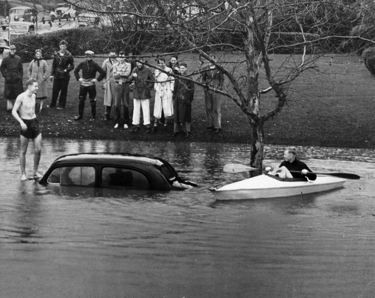 A car is submerged in water up to its windows. A man stands on the car's roof and another man paddles a kayak nearby. 