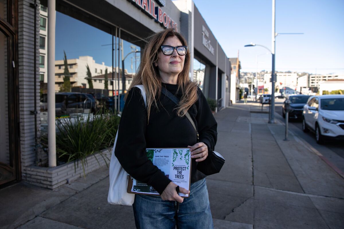 A woman in glasses, jeans and a black sweater stands on a sidewalk holding a petition