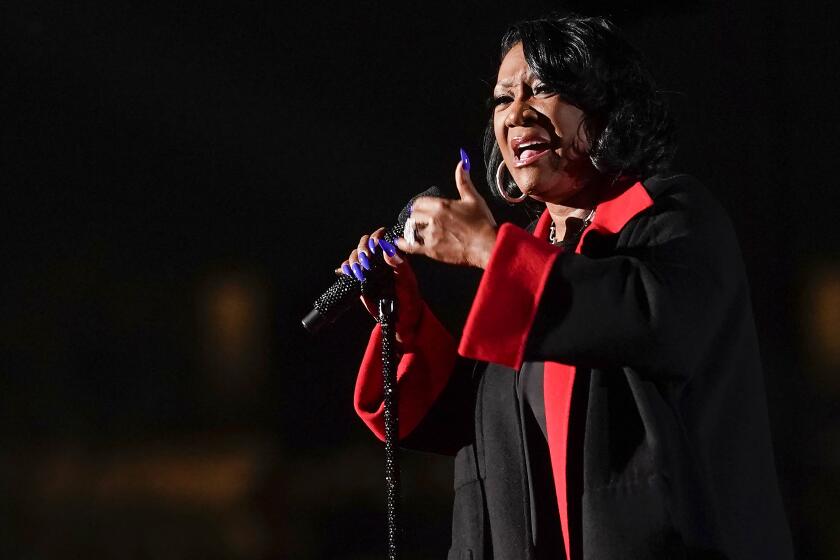 A woman with short black hair wearing a black-and-red coat and singing into a microphone