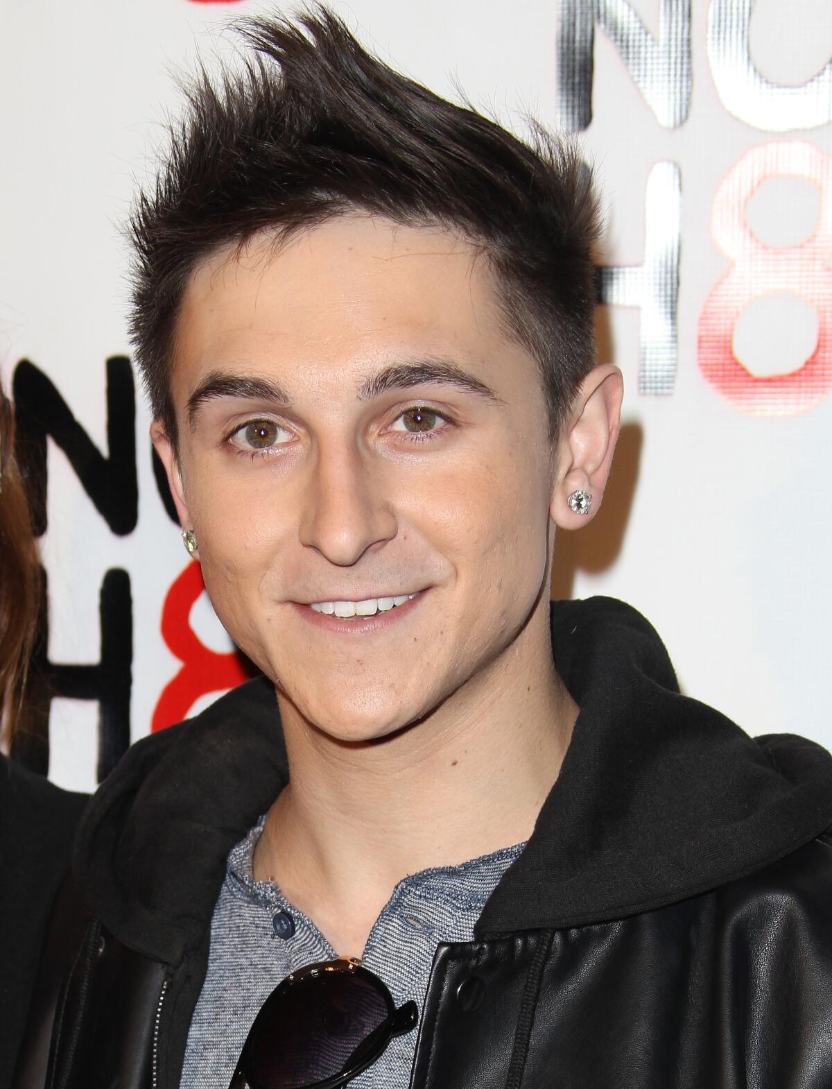 Mitchel Musso wears a black leather jacket and a gray shirt as he  stands in front of a backdrop