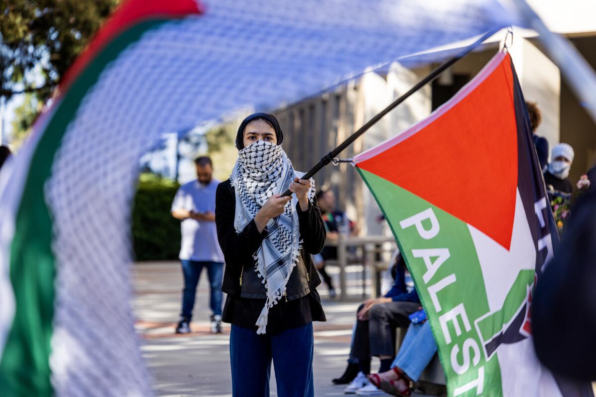 Pro-Palestinian protester framed under a supportive flag while attending a demonstration next to an encampment at UC Irvine.