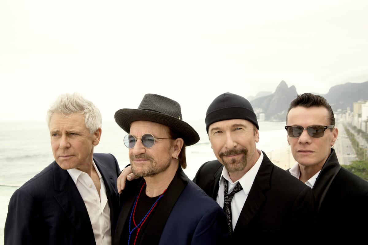 U2 members at a moody, rocky beach, all dressed in black.
