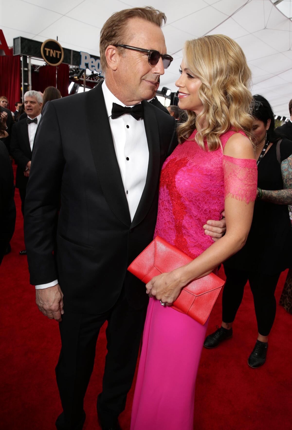 Kevin Costner wears a tuxedo and shades and smiles at Christine Baumgartner, who's wearing a pink lace gown. 
