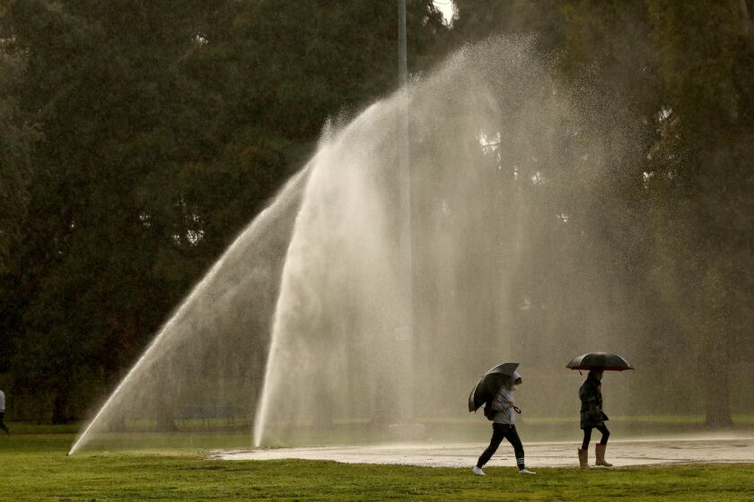 ENCINO, CA - JANUARY 29 - As rain falls a pair of people walk through spray from sprinklers heading to join a standby line in hopes of receiving the COVID-19 vaccine at the Balboa Sports Center in Encino on January 29, 2021. (Genaro Molina / Los Angeles Times)