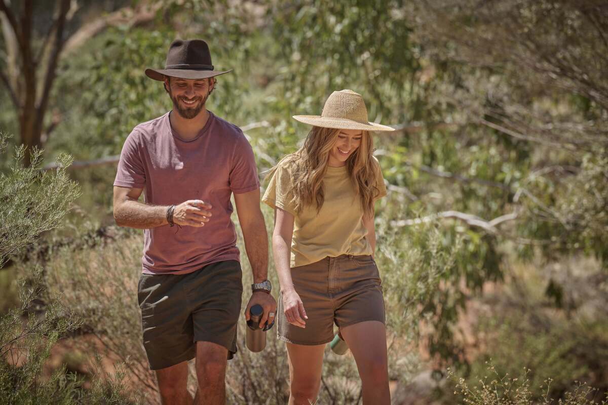 A man and a woman, in T-shirts, wide-brimmed hats and shorts, walk down a park trail.