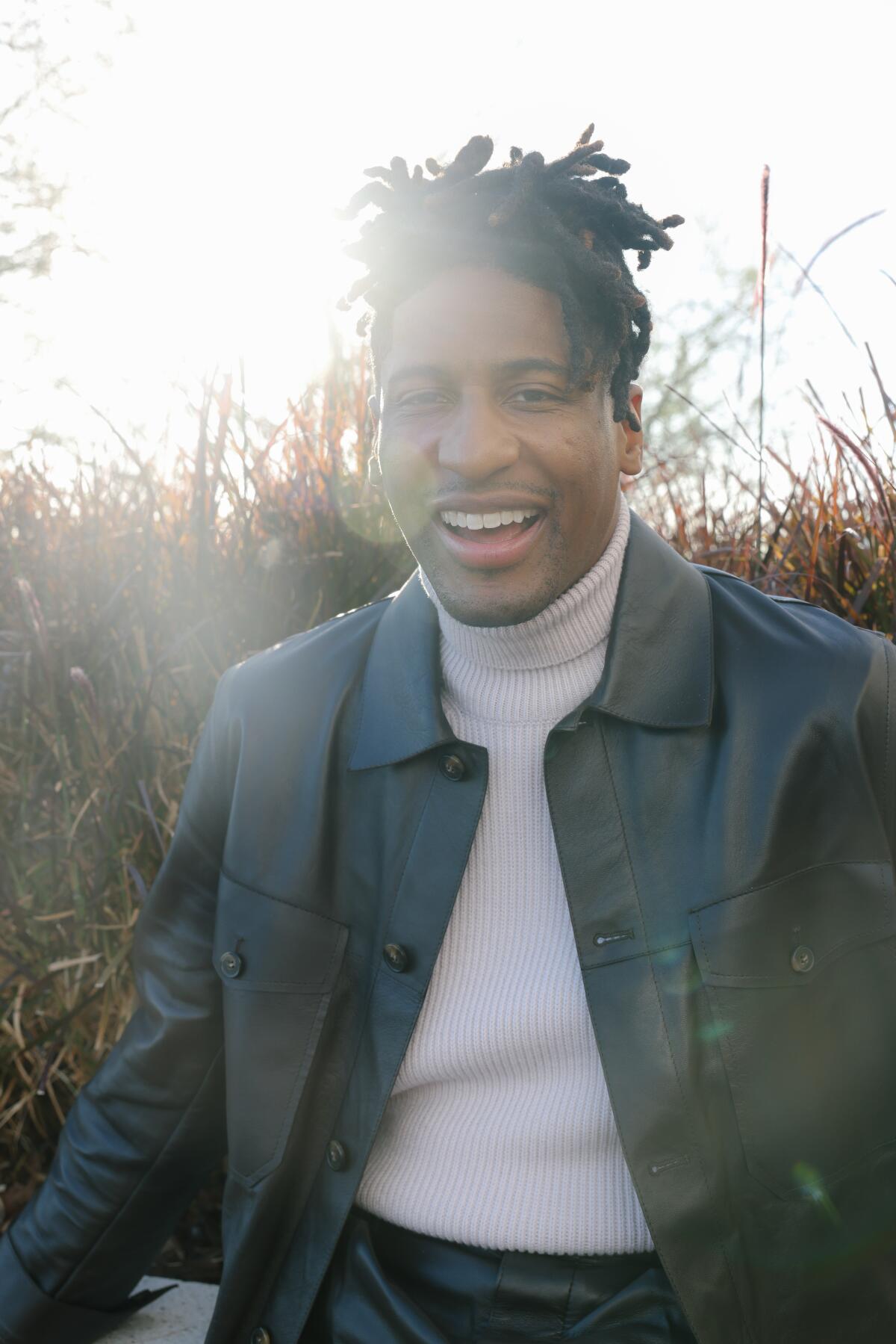 The sun shines behind the face of a smiling man with dreadlocks, a white turtleneck and a black leather jacket.