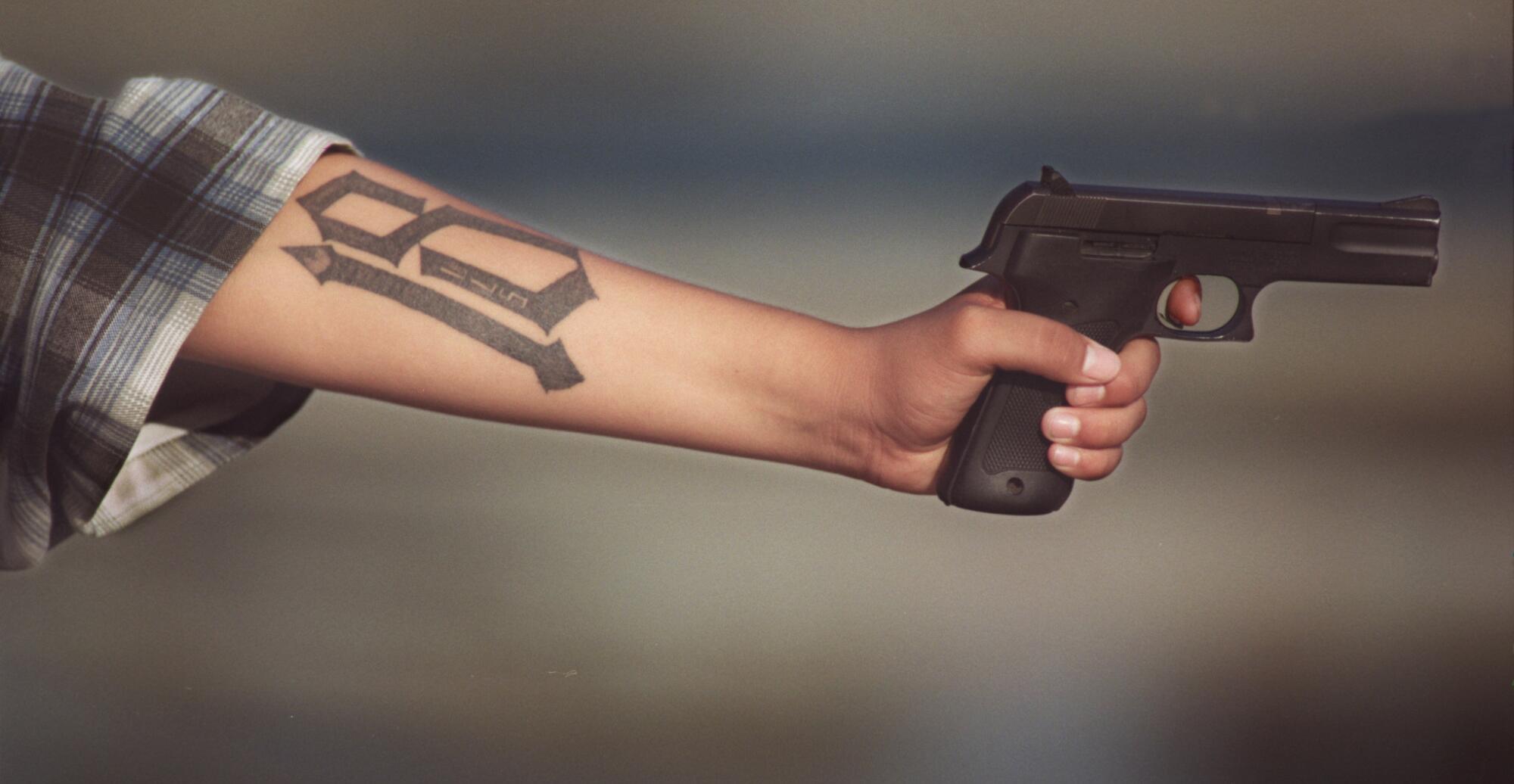From the elbow, the tattooed arm of a gang member is shown holding a gun.