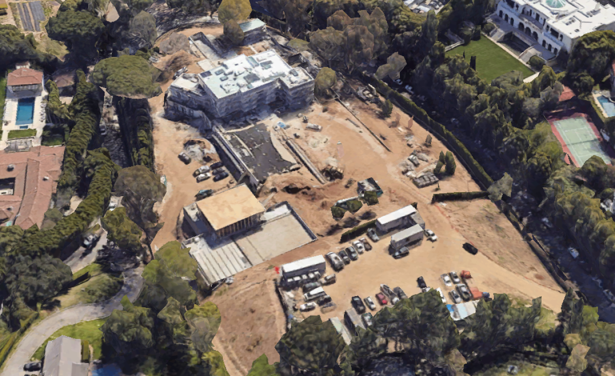 Aerial view of a mansion and other building surrounded by parked vehicles, dirt, brown grass and trees.