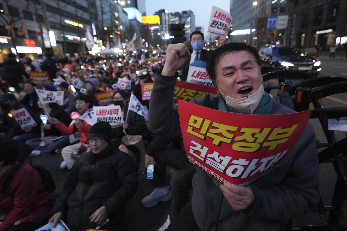 A man raises a fist as he speaks while holding a red sign, joined by a large crowd also holding signs in the street