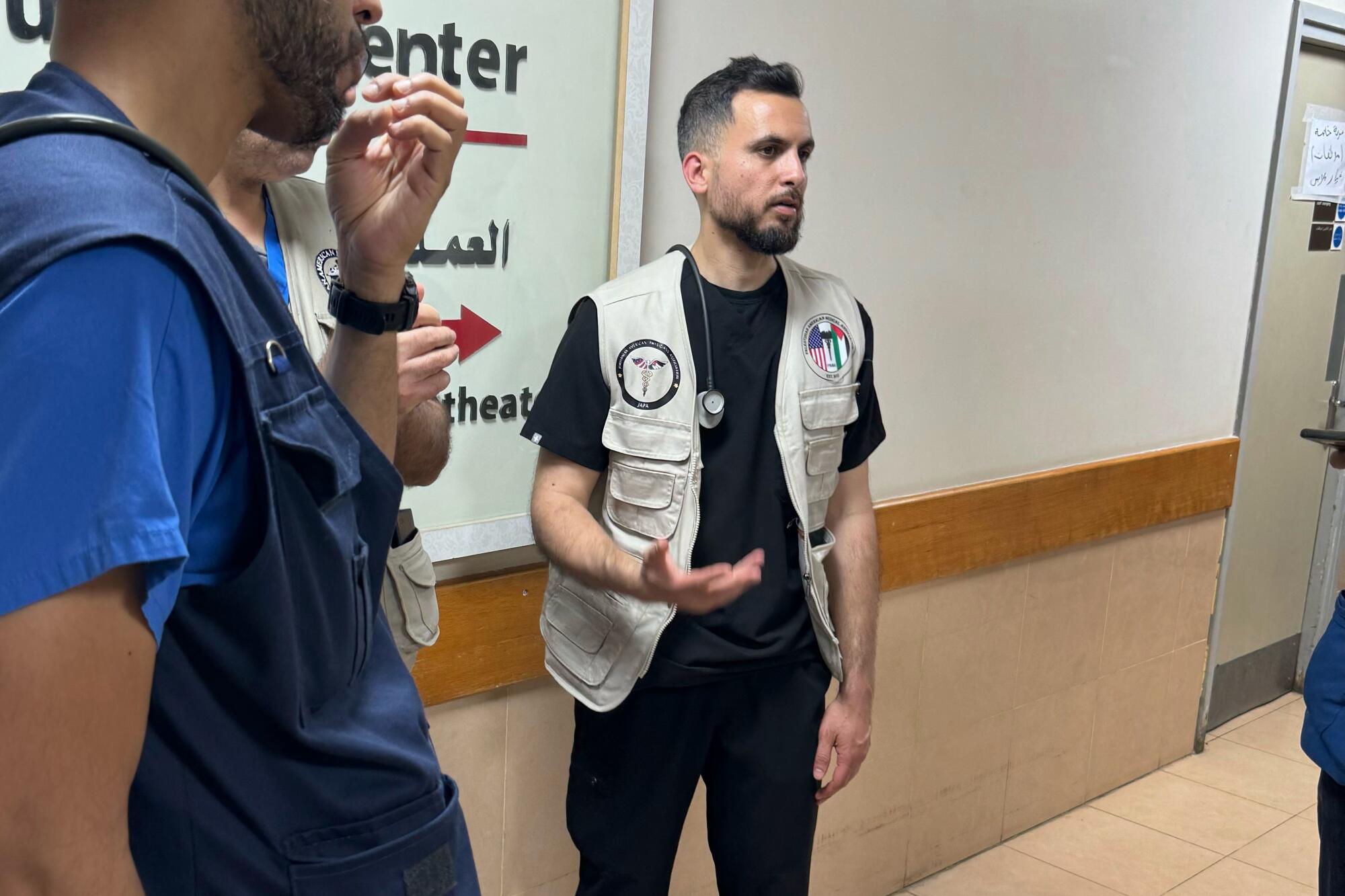 A man wearing a vest and stethoscope speaks to others in a hallway.