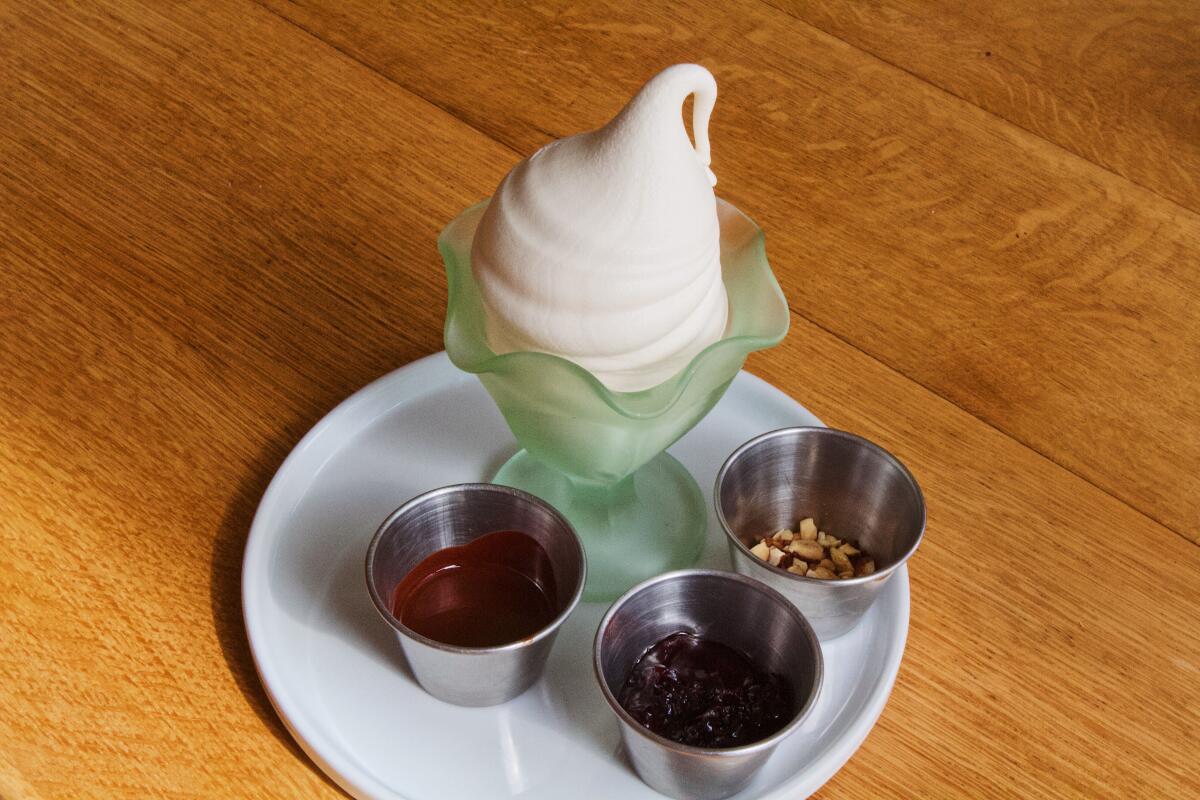 A DIY soft-serve sundae featuring vanilla soft serve in a frosted green glass. Around it are small containers of toppings.