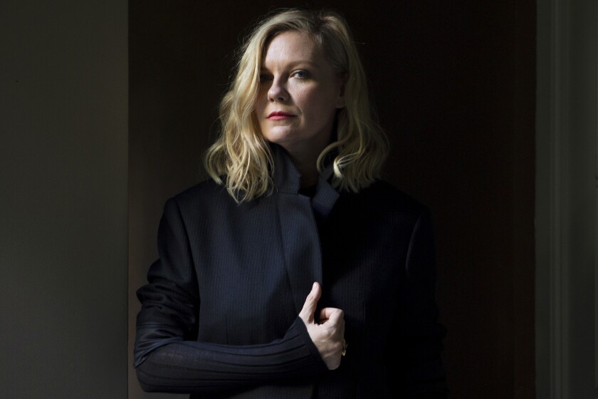 A blond woman in a dark coat poses for a portrait against a dark wall.