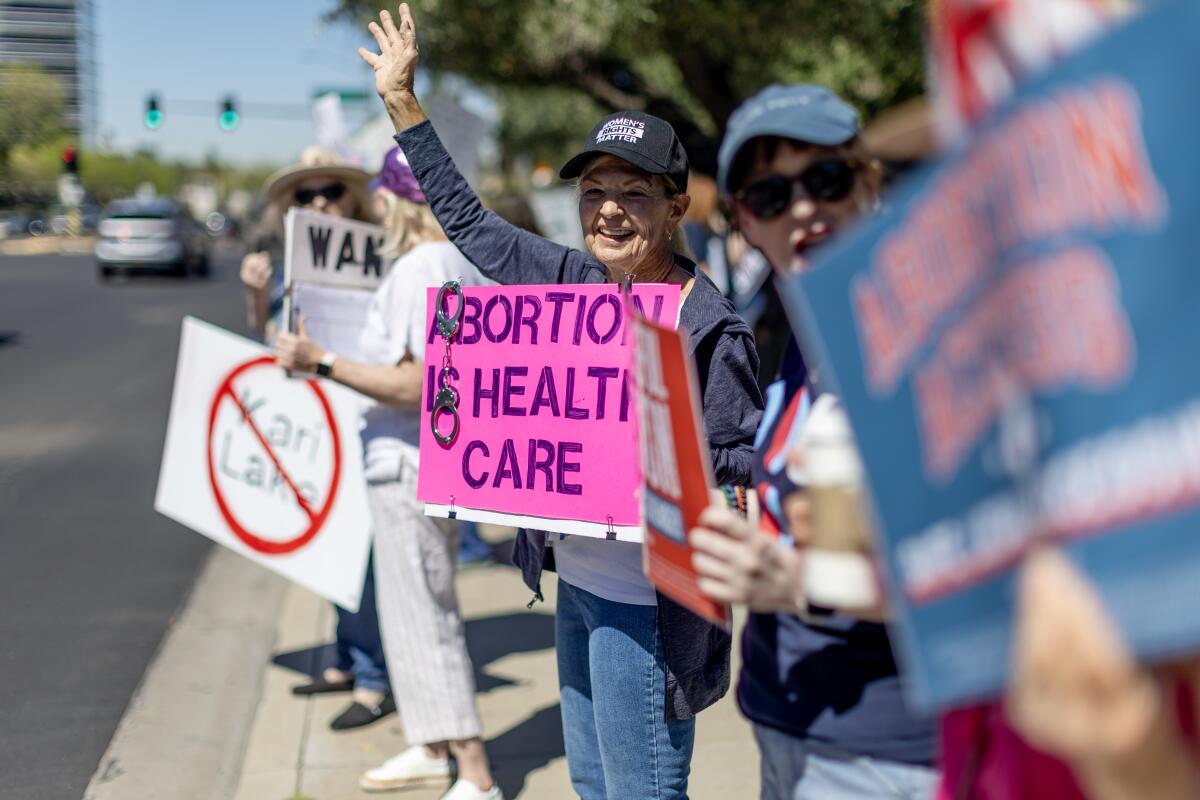 Protesters stand on a sidewalk facing the street, holding signs including a bright-pink one reading "Abortion is health care"