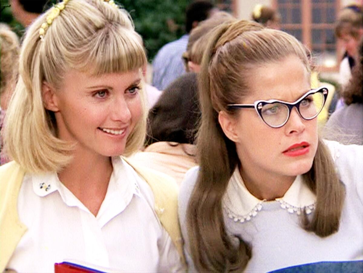 Two women looking off camera, one with short blond hair and bangs and another with long brown hair and '50s cat-eye glasses