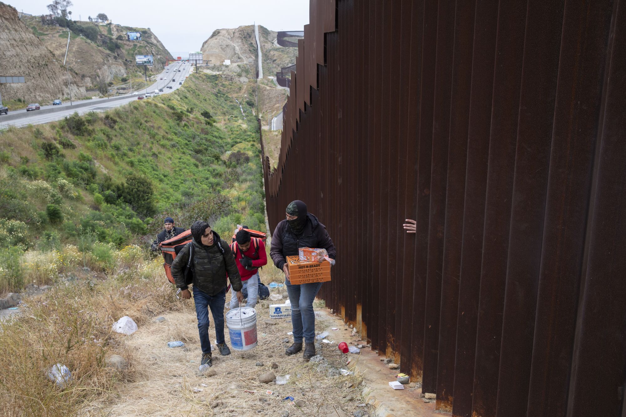 People delivering food walk a steep hill where a group of about 500-800 men traveling wait between the border walls.