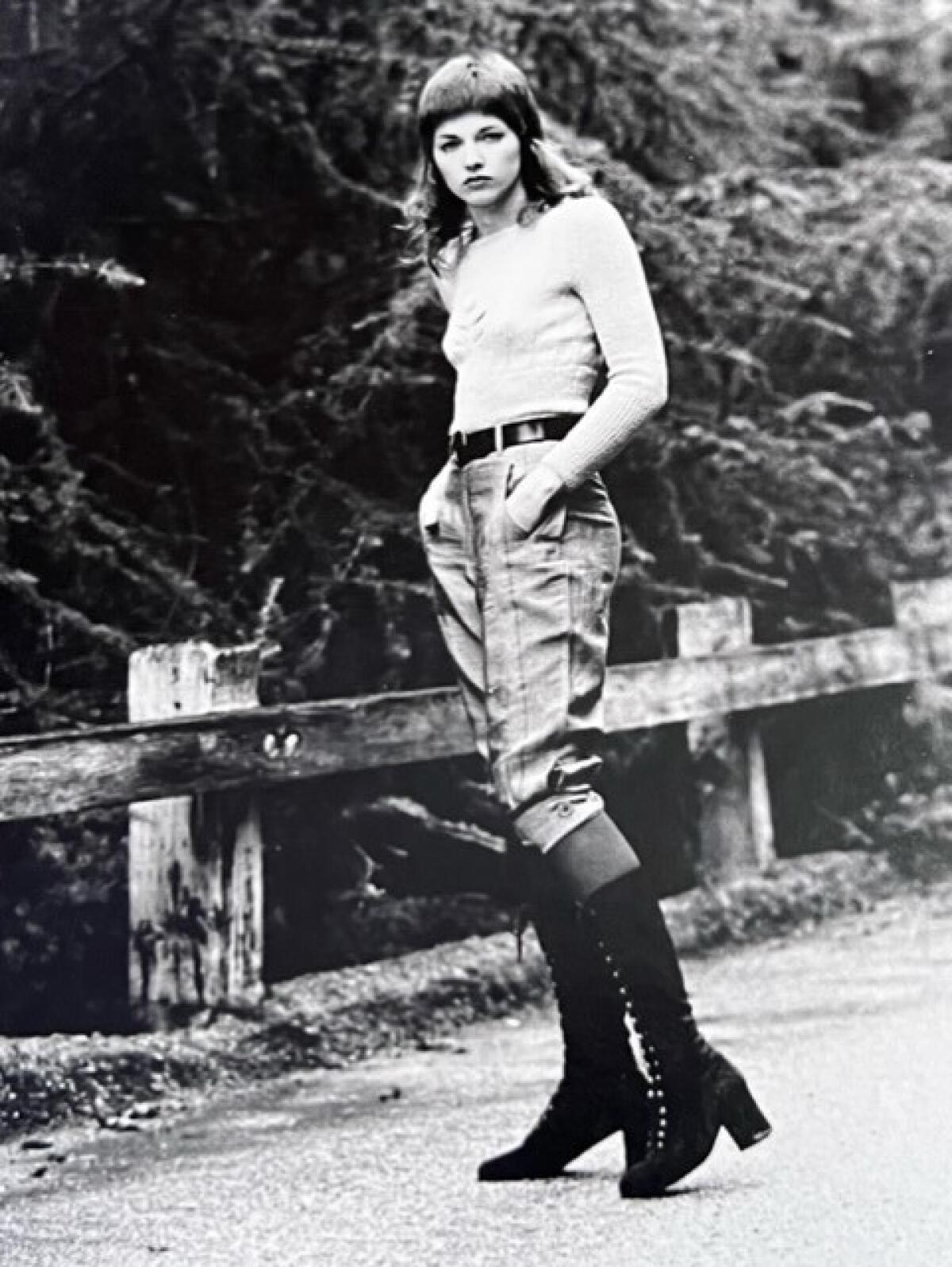 A young woman with long hair and wearing knickers and lace-up boots stands on a rural street.