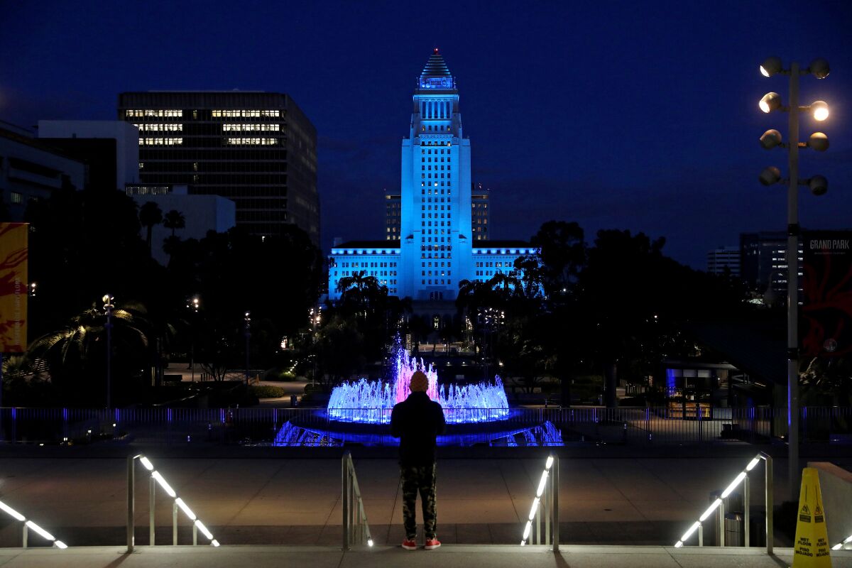 A person is silhouetted against a backdrop of a fountain and the L.A. City Hall tower lighted in blue