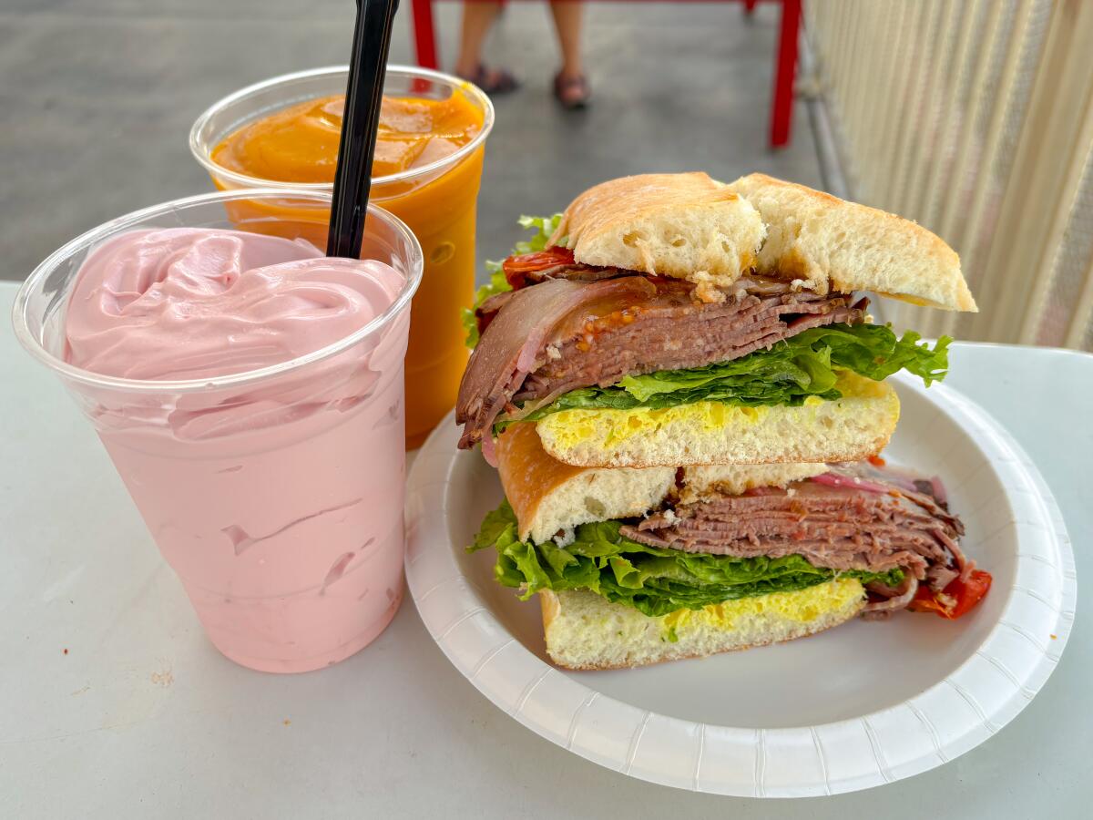 Strawberry ice cream and mango smoothie in plastic cups next to a roast beef sandwich, its halves stacked, on a paper plate