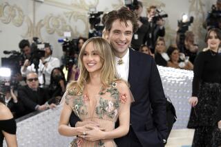 Suki Waterhouse smiles as Robert Pattinson embraces her with one arm, placing his hand along her waist