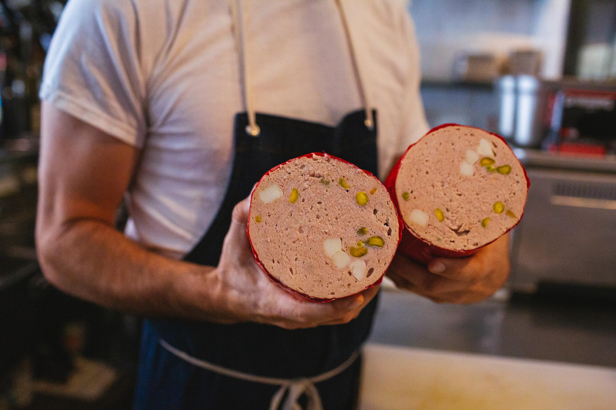 A chef in an apron holds a freshly made mortadella, sliced in half and open toward the camera.