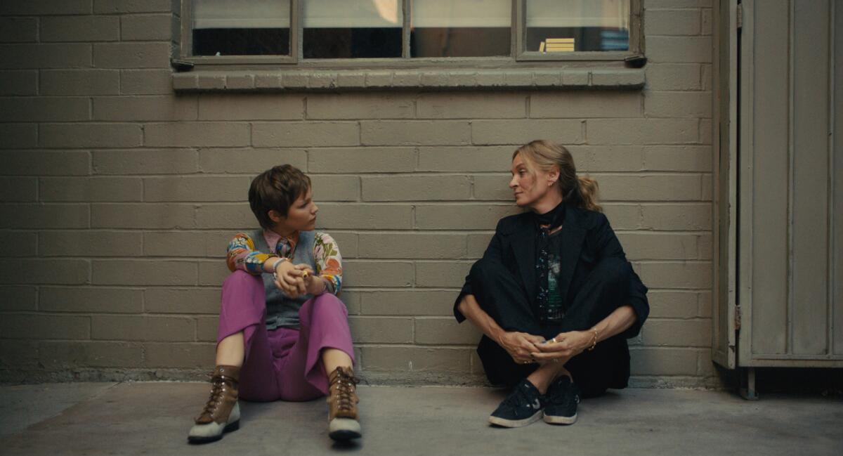 Two women talk as they sit on the ground against a drab brick wall.
