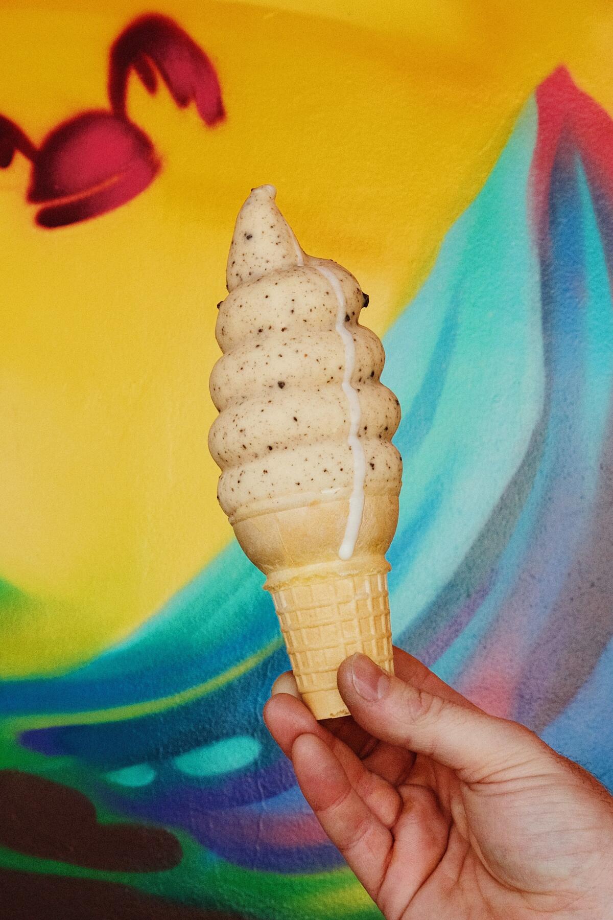 A hand holding a dripping cookies-and-cream soft serve cone in front of a rainbow mural at Heavy Handed in Studio City