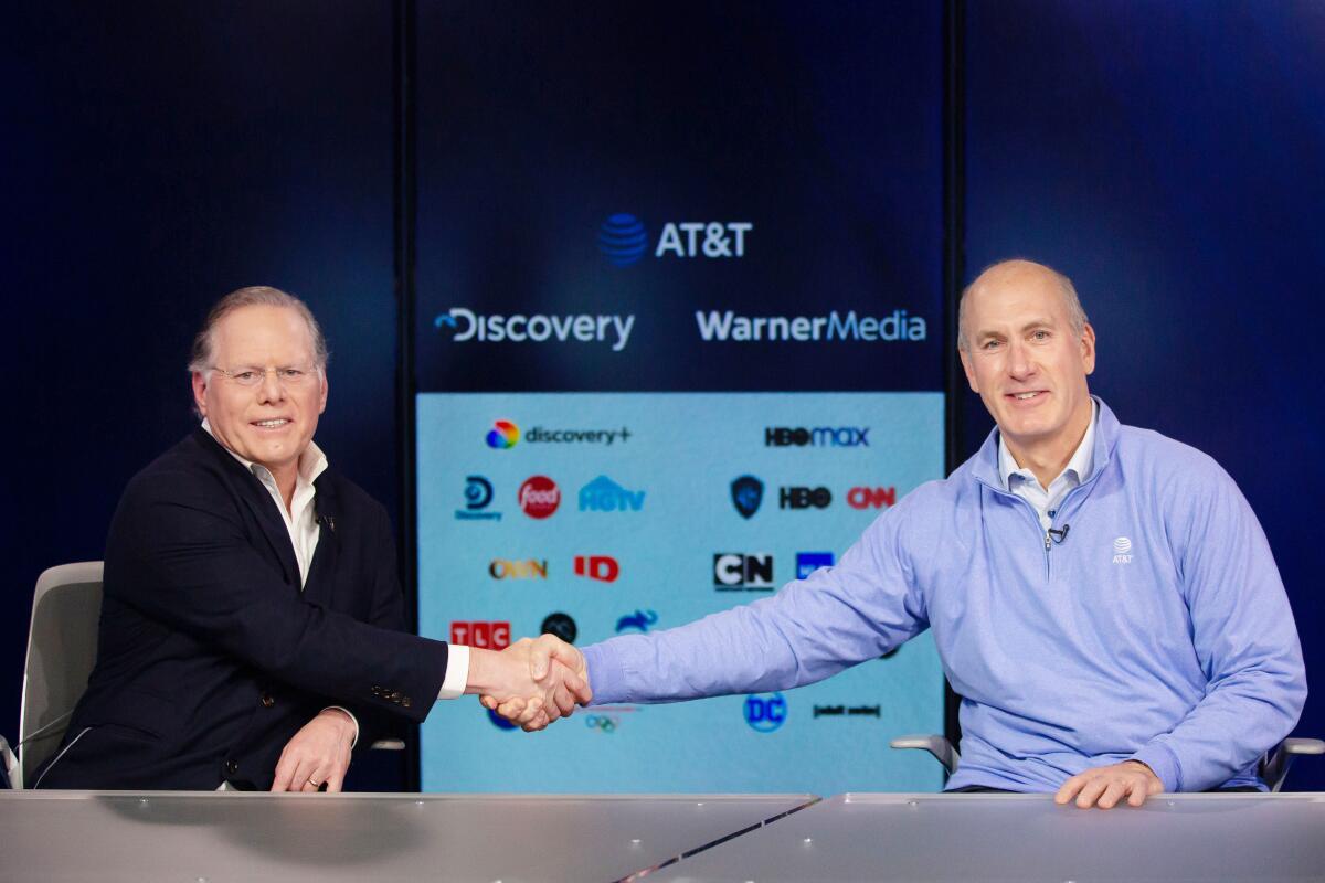 Two men shake hands in front of a screen showing logos of their companies, Discovery and WarnerMedia