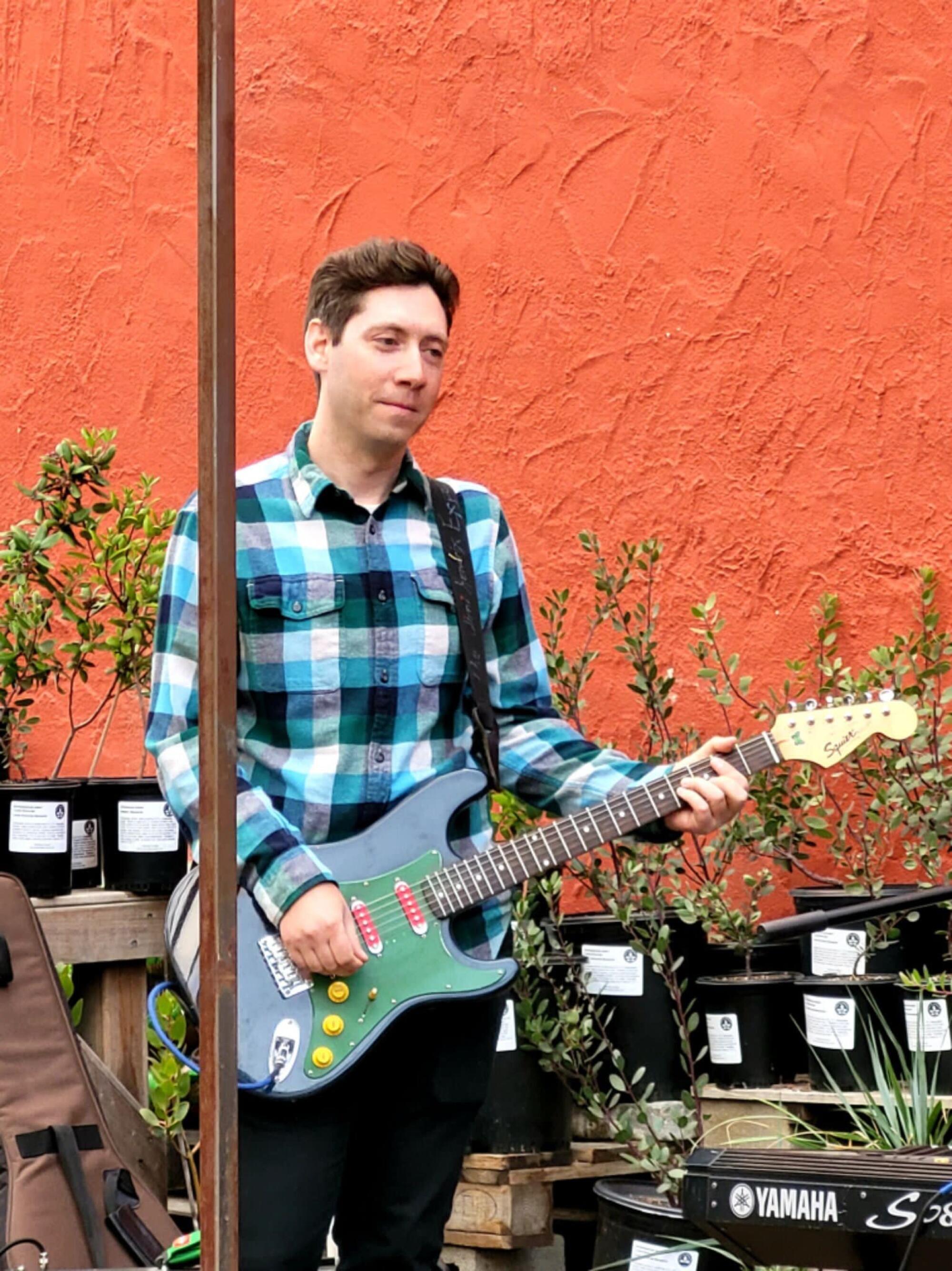 A man in a plaid shirt holding a guitar stands in front of an orange wall and potted plants