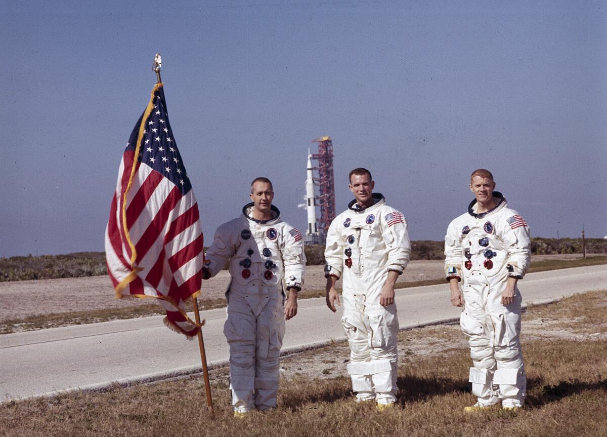 Three astronauts, one holding an American flag, stand in their spacesuits in front of a rocket on its launchpad